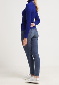 Donna in piedi di lato che indossa un maglione blu a collo alto, jeans blu aderenti e scarpe slip-on argento su uno sfondo neutro.