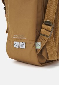 Καφέ σακίδιο φτιαγμένο από ανακυκλωμένα υλικά. Διαθέτει υφή υφάσματος, ανθεκτικά ραψίματα, ρυθμιζόμενο λουρί και ορατές ετικέτες μάρκας.