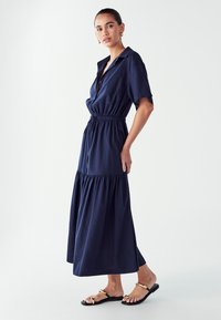 Tamsiai mėlyna trumpas rankovėmis suknelė su apykakle, elastine juosmens dalimi ir sluoksniuota sijonine dalimi. Nešiojama su plokščiomis šlepetėmis, turinčiomis minimalistinį dizainą.