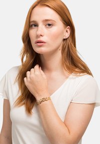 Bracciale in tono oro con collegamenti a forma di cuore e design a rete. Indossato su un braccio dalla pelle chiara; maglietta bianca, tessuto morbido con maniche trasparenti.
