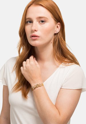 Gouden armband met hartvormige schakels en een mesh ontwerp. Draagt op een lichtgekleurde arm; witte top, zachte stof met doorschijnende mouwen.