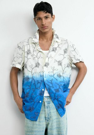 Noor mees, kes kannab lühikeste varrukatega valget särki sinise ombre ja musta lillemustriga tikandiga, helesiniseid teksapükse, seisab käed taskutes.