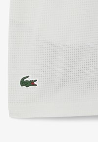 Tejido blanco texturizado con un sutil patrón de cuadrícula, con un pequeño logotipo de cocodrilo verde contorneado en blanco en la esquina inferior izquierda.