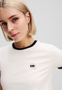 T-shirt blanc à manches courtes avec col en rib noire, présentant un design brodé d'une tête portant des lunettes de soleil et un foulard sur la poitrine.