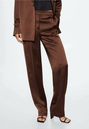 Bruine satijnen wijde broek gecombineerd met een bijpassende blazer en zwarte hakken, model met de linkerhand in de zak en de rechterhand ontspannen.