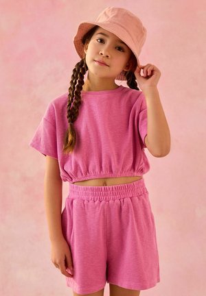 Ung pige med to fletninger, iført en pink bucket hat, pink kortærmet crop top og matchende pink shorts mod en pink baggrund.