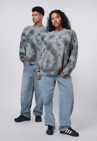 Grå sweatre med abstrakte mønstre, løs pasform og ribbede manchetter, parret med lyseblå baggy jeans og sorte sneakers.
