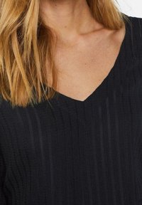 Svart topp med V-ringning, texturerat mönster med vertikala ränder och mjukt tyg. Närbild av ringning och axelparti.