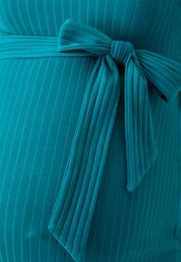 Tissu bleu sarcelle côtelé avec un détail de nœud noué, présentant un motif texturé. Le tissu tombe doucement le long du corps, mettant en valeur la silhouette.