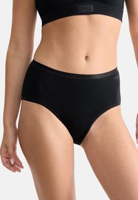 Vrouw in zwarte hoge taille slip en bijpassende zwarte sportbeha, staand tegen een effen lichte achtergrond.