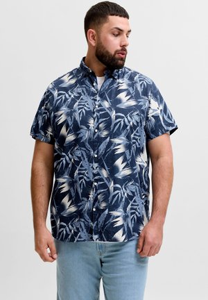 Man met kort donker haar en baard, draagend een marineblauw overhemd met korte mouwen en witte bladpatronen en lichtblauwe spijkerbroek.