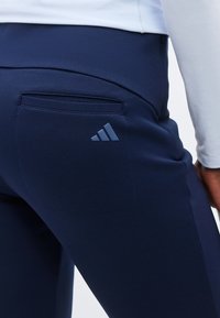 Κοντινό πλάνο ενός ατόμου που φοράει σκούρο μπλε αθλητικό παντελόνι με πίσω τσέπη και γκρι λογότυπο Adidas κοντά στην τσέπη.