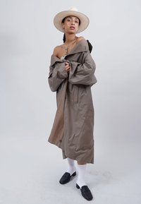 Khakifarbenen Trenchcoat mit lockerer Passform, großen Ärmeln und Seitentaschen. Kombiniert mit schwarzen Clogs und weißen gerippten Socken. Darüber ein Strohhut.