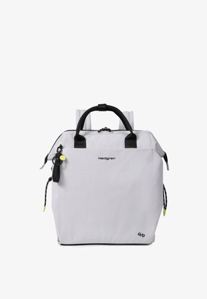Sac à dos blanc Hedgren avec poignées noires, bretelles, poche avant zippée et porte-clés noir attaché avec une étiquette jaune.