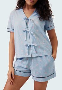 Sada pyžama ze 100% bavlny obsahuje modro-bíle pruhovanou halenu s růžovými hvězdicovými vzory, výstřihem ve tvaru V a mašličkami, doplněnou o odpovídající šortky s černým lemem.