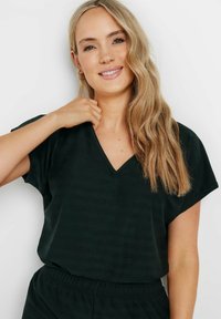 Top de color verde oscuro con escote en V y mangas cortas, que presenta una textura suave y un sutil patrón de rayas. La modelo está ajustando el escote.