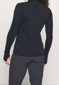 Svart långärmad topp med hög krage, figurnära design och texturerade accenter. Kombinerad med mörkgrå byxor som visar en strömlinjeformad silhuett.