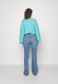 Pull turquoise à manches longues associé à un jean bleu évasé. Le pull a un col rond ; le jean possède des poches arrière et une étiquette.