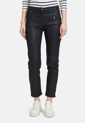 Pantalons noirs ajustés en matériau brillant, dotés de poches zippées et d'une fermeture à bouton, associés à des baskets blanches.