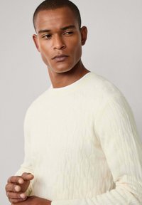 Suéter de punto color crema claro con un patrón de diamante texturizado. Presenta un cuello acanalado y mangas largas.