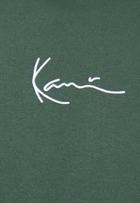 Tessuto verde con una texture a righe presenta un logo ricamato in bianco. Il design è minimalista, senza ulteriori motivi o accenti.