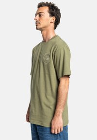 Olive green t-shirt with a crew neck and short sleeves. Features a small embroidered logo on the left chest. Cotton material, lightweight texture.