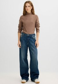 Suéter de cuello alto marrón, textura suave, combinado con jeans de mezclilla azul sueltos, de cintura alta y pierna ancha, complementados con zapatillas blancas.