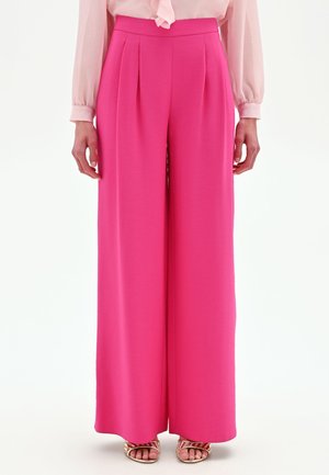 Femme portant un pantalon large taille haute rose vif, un chemisier rose clair et des sandales ouvertes dorées, debout devant un fond blanc.