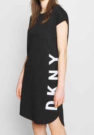 Kobieta w czarnej sukience z krótkim rękawem, kieszenią na piersi i białym pionowym logo "DKNY" z boku na jednolitym tle.