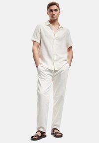 Lys beige kortærmet skjorte med knapper og et struktureret mønster, parret med hvide bukser og brune sandaler. Enkel og moderne design.