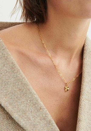 Guld kædehalskæde med en lille firkløvervedhæng båret om en kvindes hals, iført en beige tekstureret blazer.