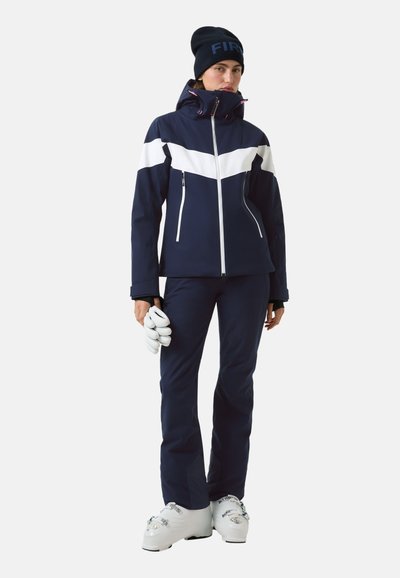 Granatowa kurtka narciarska z białym wzorem w kształcie szewrona, kieszeniami na suwak i wysokim kołnierzem. W komplecie granatowe spodnie narciarskie i białe buty narciarskie. W rękach białe rękawice.