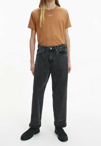 Hnědé tričko s krátkým rukávem s logem "Calvin Klein Jeans", v kombinaci s černými vysokými džíny a černými teniskami. Modelka stojí vzpřímeně.