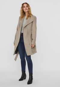 Beige kappa med draperad krage, långa ärmar och dragsko i midjan. Matchad med mörka jeans och svarta ankellånga stövlar, med en slät textur.