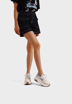 Persona con pantalones cortos negros, camiseta negra con gráfico y zapatillas blancas con detalles de estampado de leopardo, de pie frente a un fondo claro y sencillo.