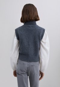 Person med kort brunt hår iført en ærmeløs grå strikvest over en hvid langærmet bluse og grå bukser, som vender væk.