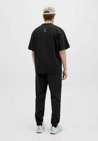 Camiseta negra de gran tamaño con mangas cortas, que presenta una impresión de logo en la parte posterior. Combinada con pantalones jogger negros y zapatillas blancas.