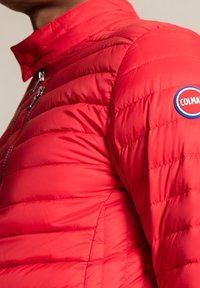 Veste matelassée rouge avec un col montant, une texture matelassée, une fermeture éclair argentée et un logo circulaire bleu et rouge sur l'épaule.