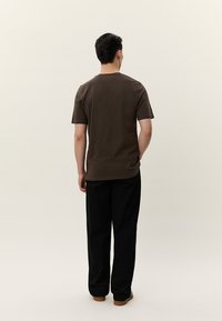 Man som står med ryggen mot kameran, klädd i en mörkbrun kortärmad skjorta, svarta byxor och bruna skor mot en neutral bakgrund.