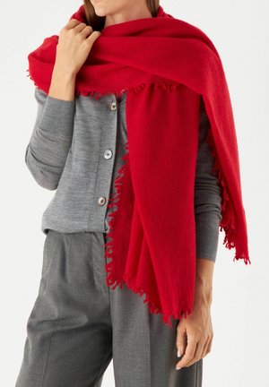 Rode wollen sjaal met franjes, gewikkeld om de schouders over een grijze cardigan, gekenmerkt door een zachte textuur en een levendige kleur.