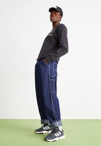 Mörkgrå sweatshirt med broderad logotyp, lösa blå denimbyxor med kontrasterande sömmar och svart keps. Sportiga låga sneakers.