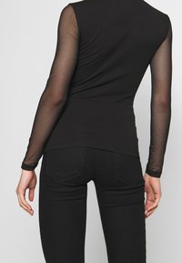 Svart topp med långa genomskinliga ärmar och en figursydd design, med ett slätt tyg och rundad halsringning, parat med svarta jeans.