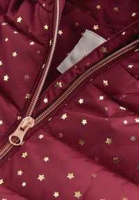 Chaleco acolchado burdeos con patrones de estrellas doradas, cremallera frontal con herrajes en oro rosa y una textura suave y brillante.