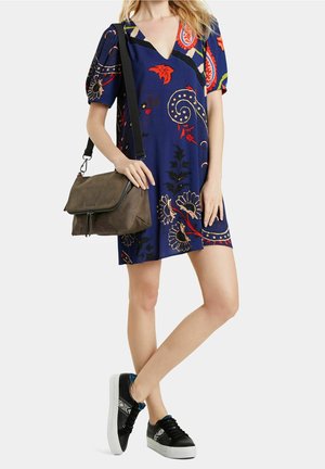 Femme portant une robe courte bleue à fleurs, des baskets noires aux semelles blanches, et un sac bandoulière marron avec une sangle noire.