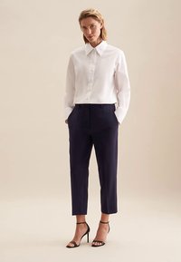 Camicia bianca con bottoni e colletto, abbinata a pantaloni sartoriali blu navy e sandali neri con cinturino, che mostrano un design pulito e fresco.