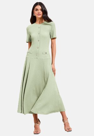 Kvinde iført tekstureret lys grøn knappet kjole med korte ærmer og lommer, kombineret med beige sandaler med høj hæl og stropper, stående mod en hvid baggrund.