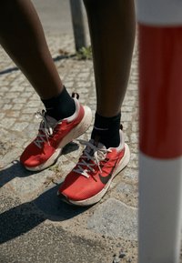 Scarpe da ginnastica rosse con tomaia in tessuto testurizzato, suole bianche e dettagli neri, indossate con calzini neri. Lo sfondo presenta un pavimento in ciottoli.