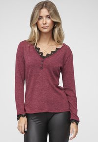 Weinrotes Langarmshirt mit V-Ausschnitt, das schwarze Spitzenbesätze am Ausschnitt und den Bündchen sowie drei dekorative Knöpfe auf der Vorderseite aufweist.