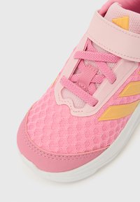 Pantof sport roz, cu partea superioară din plasă, închidere cu șiret și velcro și accente aurii. Talpă moale din cauciuc cu un model texturat.