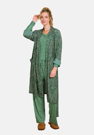 Groene lange cardigan met bloemenpatroon en zakken, gecombineerd met een groen-wit gestreept topje en bijpassende broek. Accessoires zijn bruine slippers.